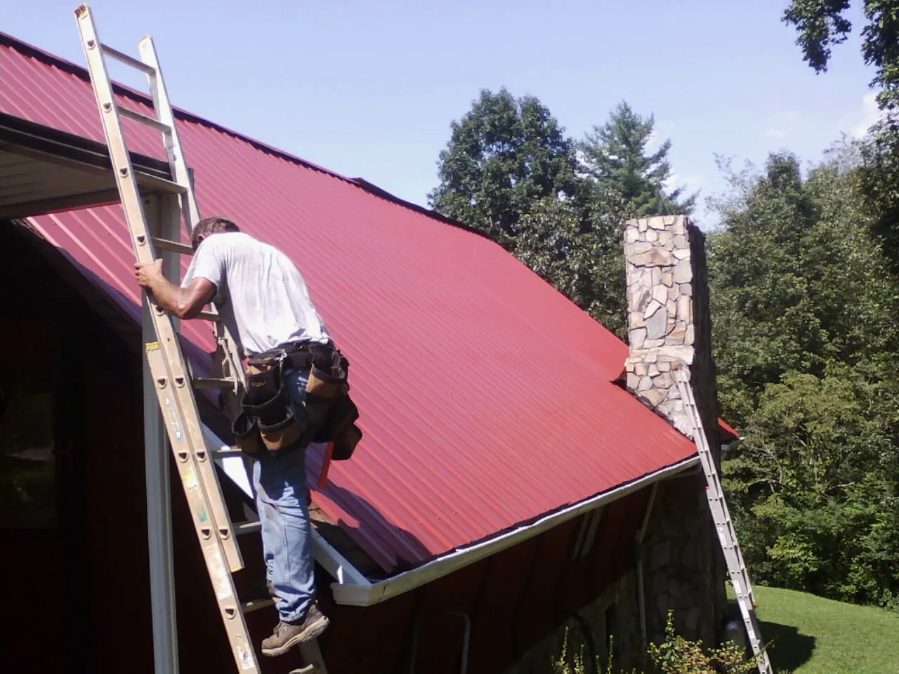 Roofer on a ladder beside a red metal roof and stone chimney — Soffit & Fascia Repair by Roofing Brothers in Henryville